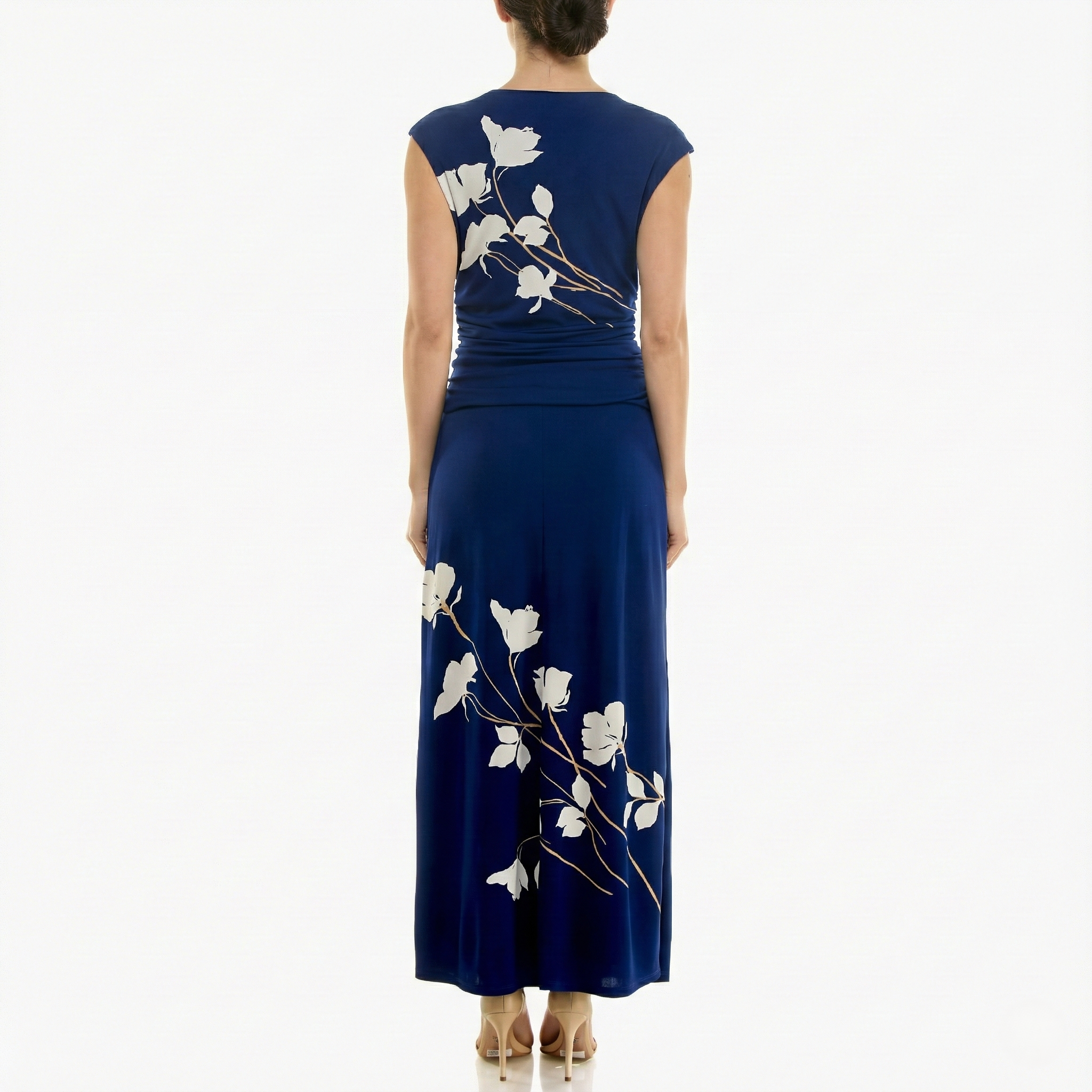 Back view of a women’s navy blue sleeveless maxi dress with ivory floral branch print, fitted ruched bodice, flowing skirt, and ankle-length hem, shown with hair in a low bun and nude heels.