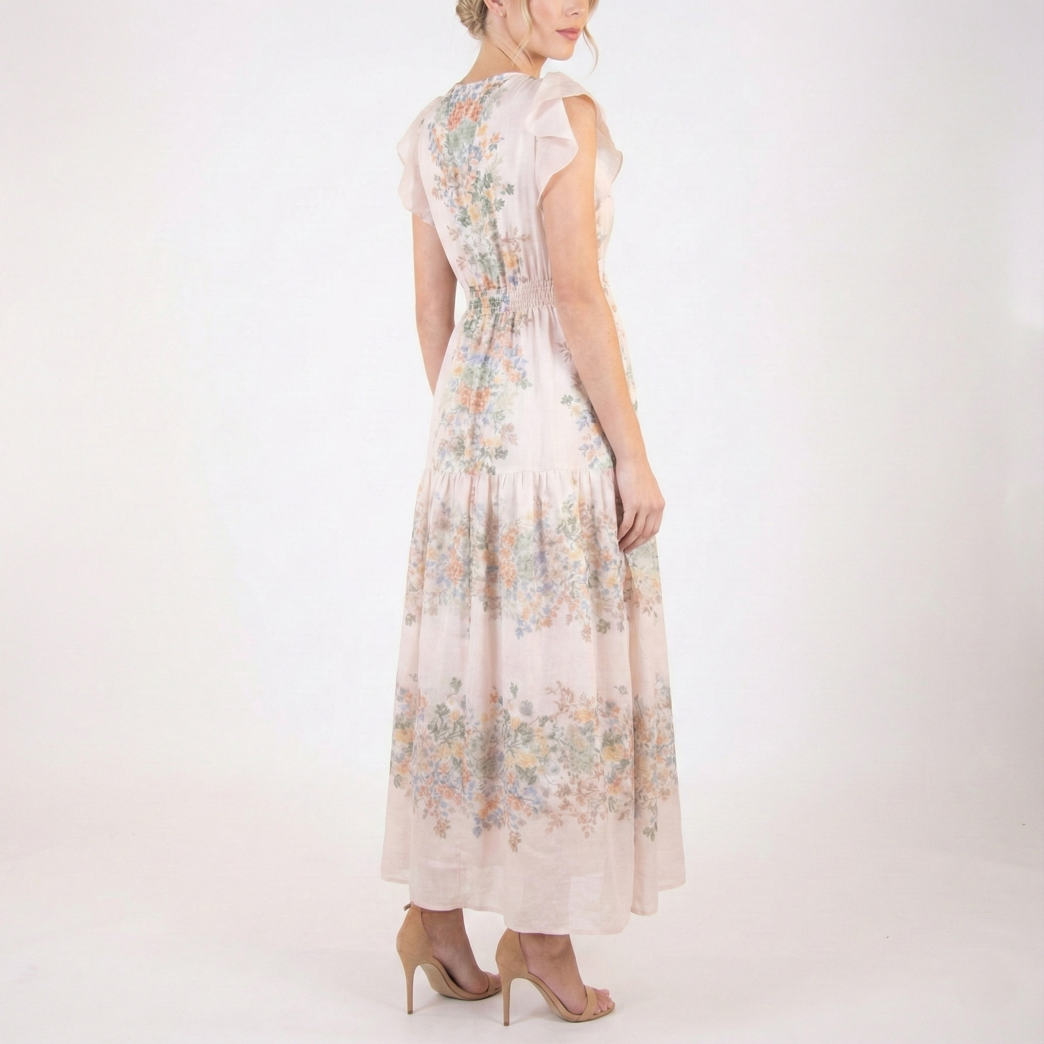 Back angled view of blush pink floral chiffon maxi dress with flutter sleeves, smocked waist, and flowing tiered skirt, styled with nude heels.