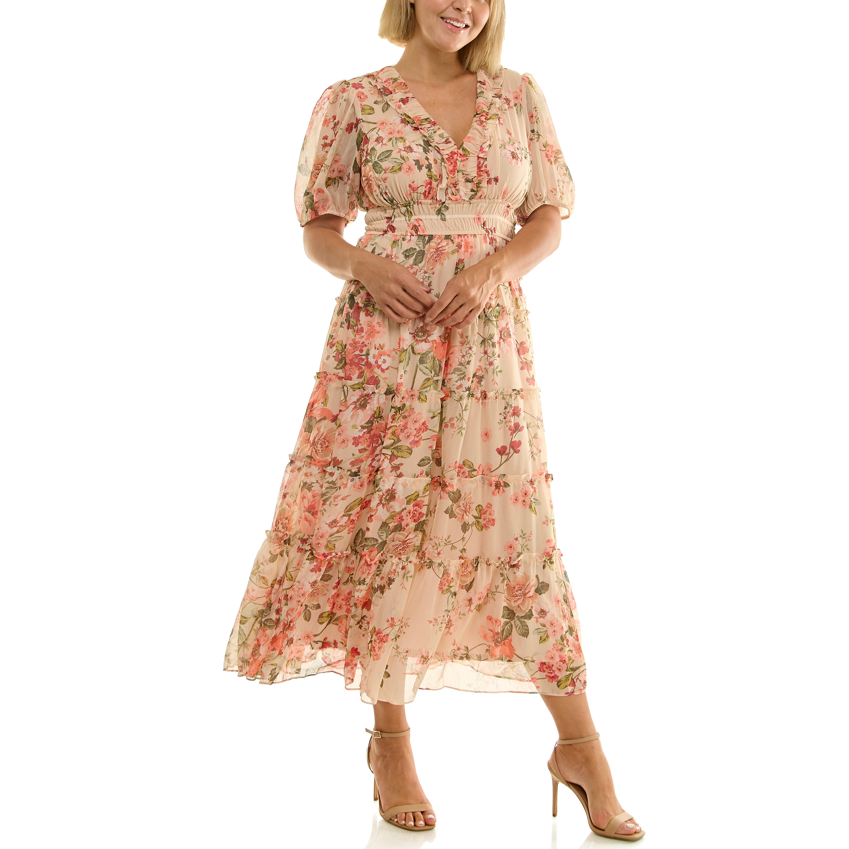 Front view of a woman wearing a light pink floral midi dress with a V-neck ruffle detail, short puff sleeves, elastic waist, and flowing tiered skirt, standing in nude high-heeled sandals against a white background.