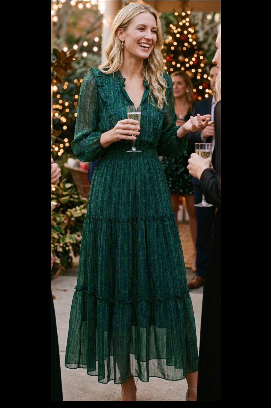 A woman wearing a dark green tiered tulle midi dress with sheer long sleeves, holding a champagne flute in a festive indoor party setting with blurred Christmas trees and lights in the background.
