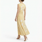 Back view of cornsilk yellow sleeveless midi dress with white floral print, softly ruched waist, flowing skirt, and nude high-heel sandals.