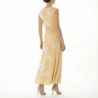 Back angled view of a cornsilk yellow sleeveless midi dress with soft white floral print, ruched waist detail, flowing skirt, and nude high-heel sandals.