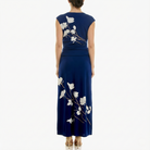 Back view of a women’s navy blue sleeveless maxi dress with ivory floral branch print, fitted ruched bodice, flowing skirt, and ankle-length hem, shown with hair in a low bun and nude heels.