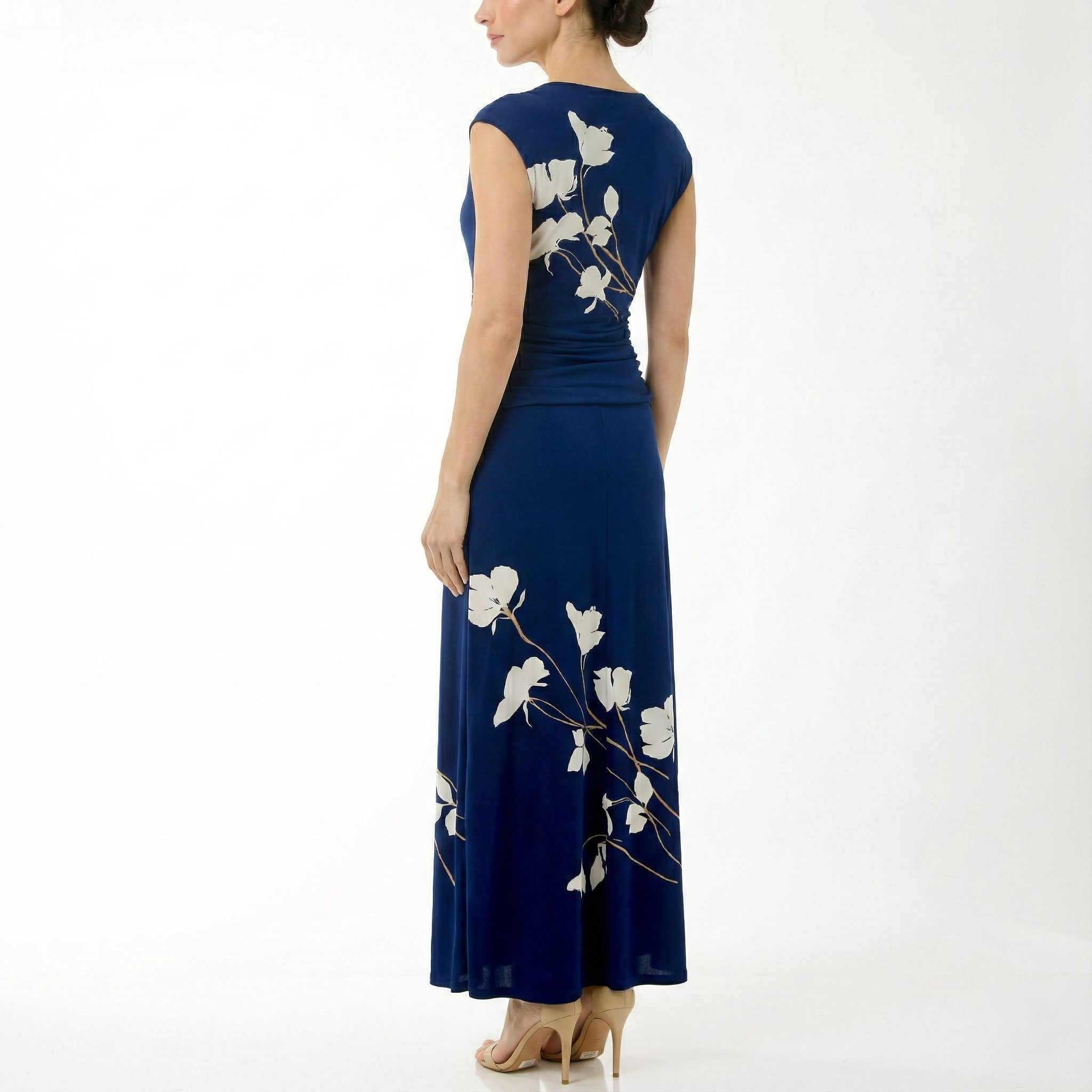 Back angled view of a navy sleeveless midi dress with ivory floral print and tan branch detail, ruched waist, flowing skirt, and nude high-heel sandals.