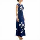Side view of a women’s navy blue sleeveless maxi dress featuring ivory floral branch print, ruched waist, round neckline, and flowing ankle-length skirt, worn with nude high-heel sandals.