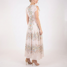 Back angled view of blush pink floral chiffon maxi dress with flutter sleeves, smocked waist, and flowing tiered skirt, styled with nude heels.