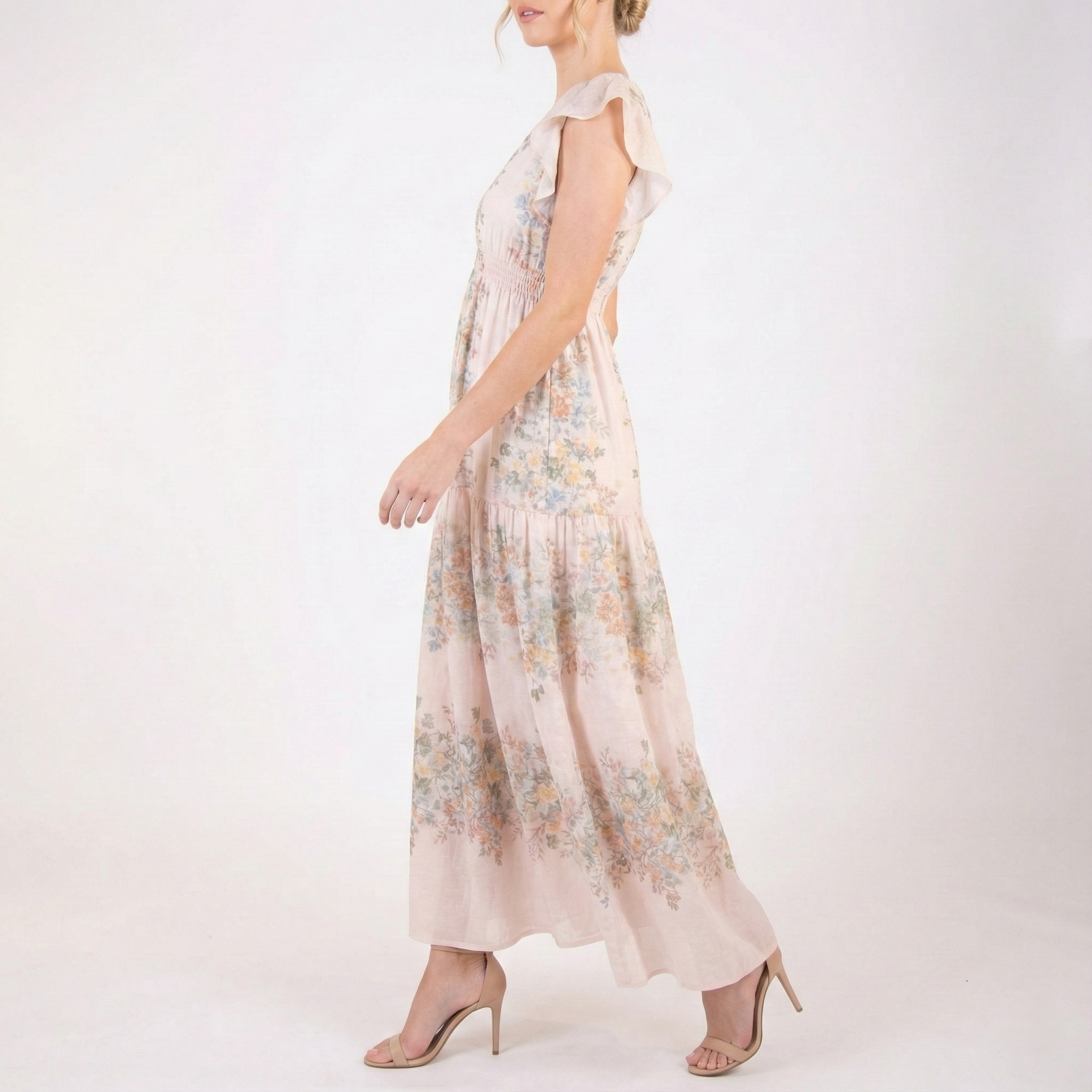 Side view of blush pink floral chiffon maxi dress featuring flutter sleeves, smocked waist, flowing tiered skirt, and lightweight sheer fabric, styled with nude heels.