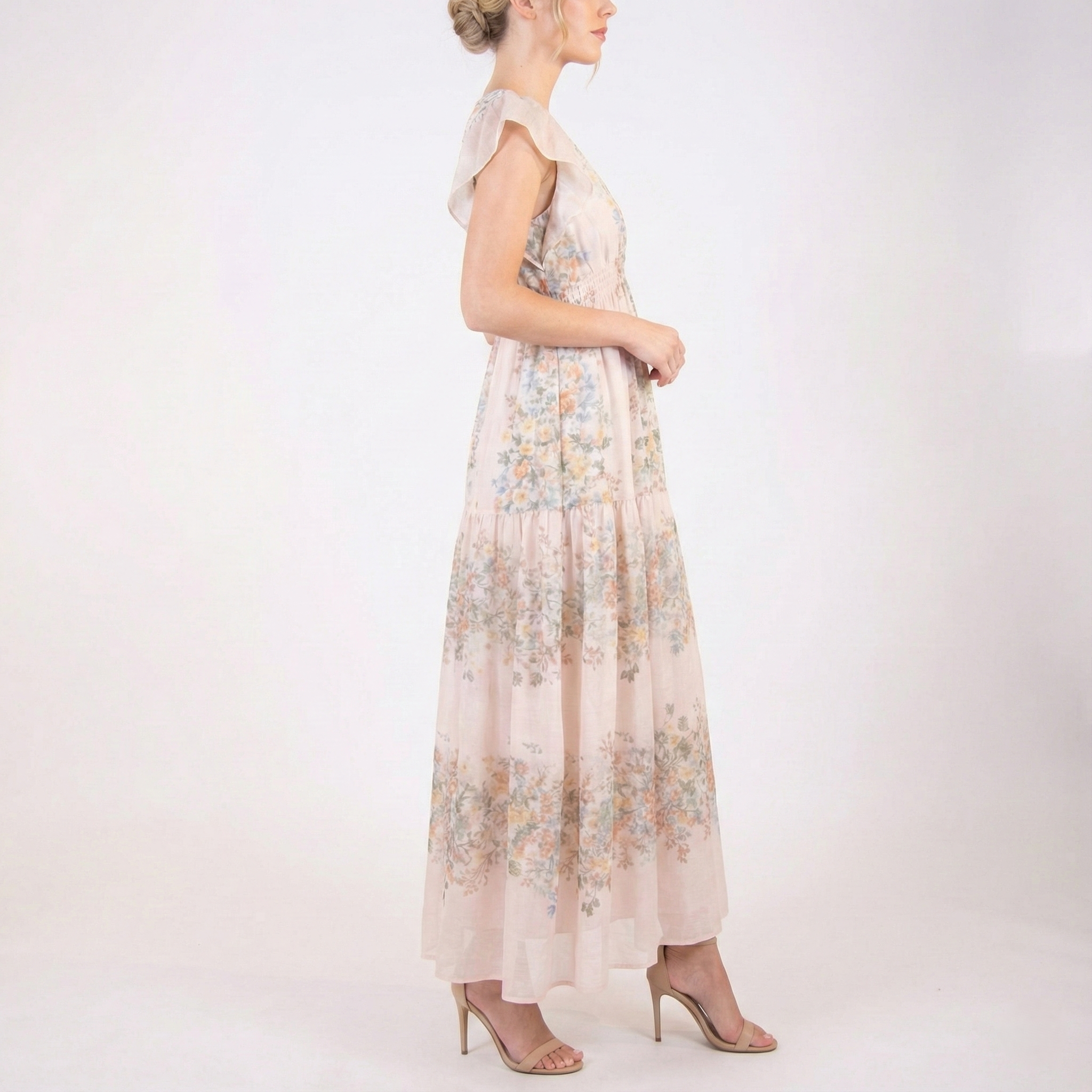 Side profile of blush pink floral chiffon maxi dress with flutter sleeves, smocked waist, lightweight sheer fabric, and tiered skirt, worn with nude heels.