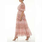 Side view of model walking in blush pink floral chiffon maxi dress featuring short puff sleeves, V-neck bodice, fitted waist, and airy tiered skirt.