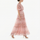 Side profile of model walking in blush pink floral chiffon maxi dress with puff sleeves, V-neck bodice, fitted waist, and flowing tiered skirt.