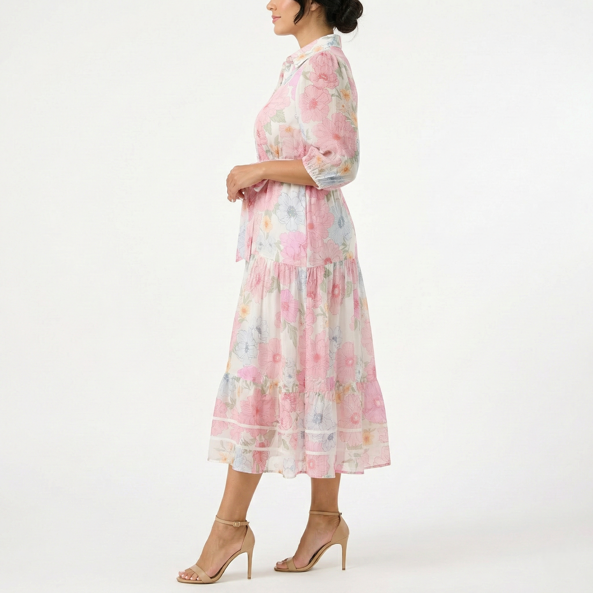 Side profile of a model wearing a sheer pastel floral midi dress with elbow-length sleeves, a tiered skirt, and a waist tie, paired with nude high-heel sandals