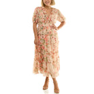 Front view of a smiling woman wearing a light pink floral midi dress with a V-neckline, ruffled trim, short puff sleeves, elastic waist, and tiered skirt, paired with nude high-heeled sandals on a white background.